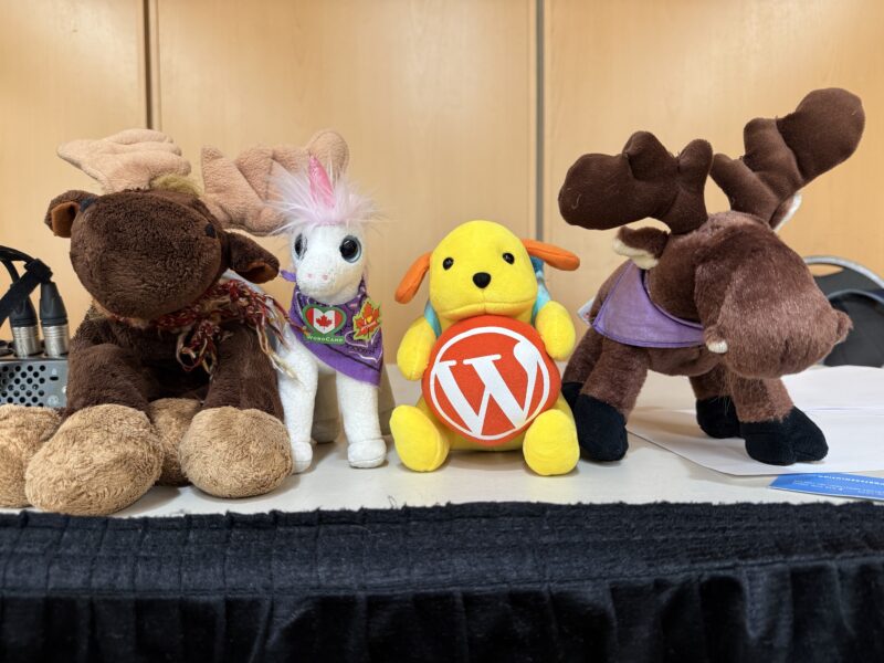 From left to right, a stuffed moose, a stuffed unicorn with Canada charms (a Canadian flag inside a heart and a maple leaf), a Wapuu with an orange WordPress logo and another moose standing next to each other on a table at WordCamp Canada.